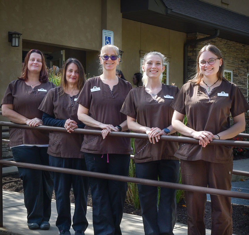 Five people wearing brown medical scrubs stand side by side outdoors in front of a building. They are all smiling and are positioned behind a railing. The background includes a sign with a wheelchair symbol and some landscaping with bushes and plants, hinting at their work as veterinarians.