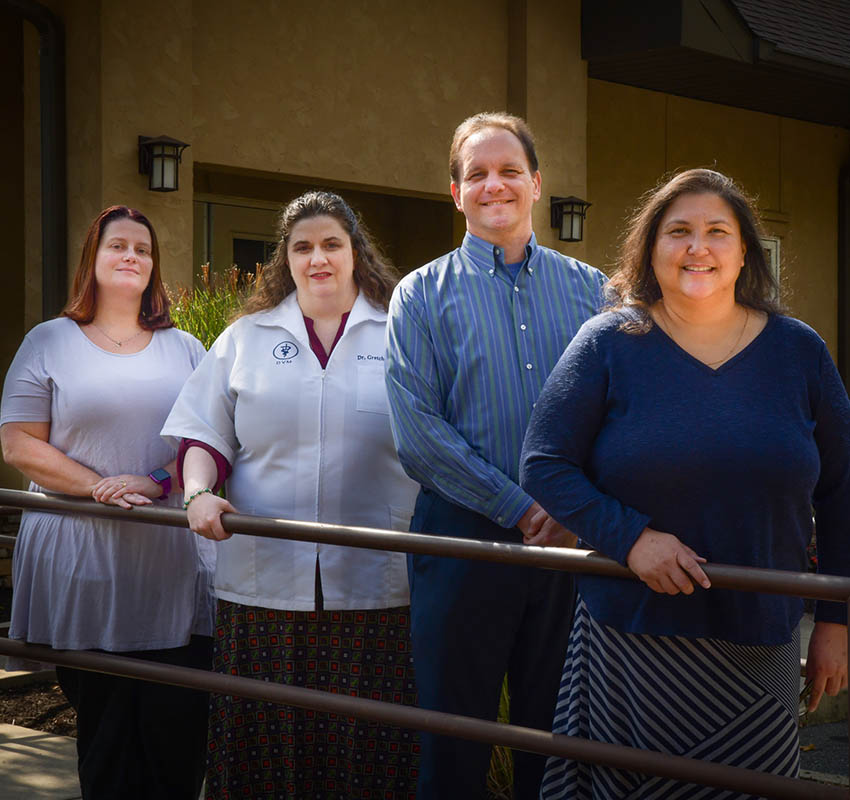 Four people stand outdoors near a handrail and a building, posing for a group photo. The group consists of three women and one man. They are casually dressed, except for one woman wearing a white medical coat, indicating she might be a veterinarian. The background features greenery and part of the building.
