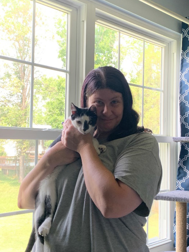 A woman with dark hair, wearing a grey shirt, stands in front of large windows holding a white and black cat. The windows show a view of a green lawn and trees. The veterinarian's cat looks towards the camera.