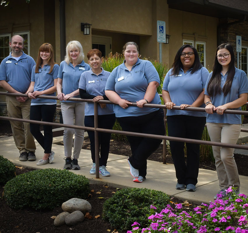 A group of seven individuals wearing blue polo shirts stand in a line, smiling, in front of a building with landscaping that includes bushes and pink flowers. They appear to be veterinary staff members posing for a group photo.