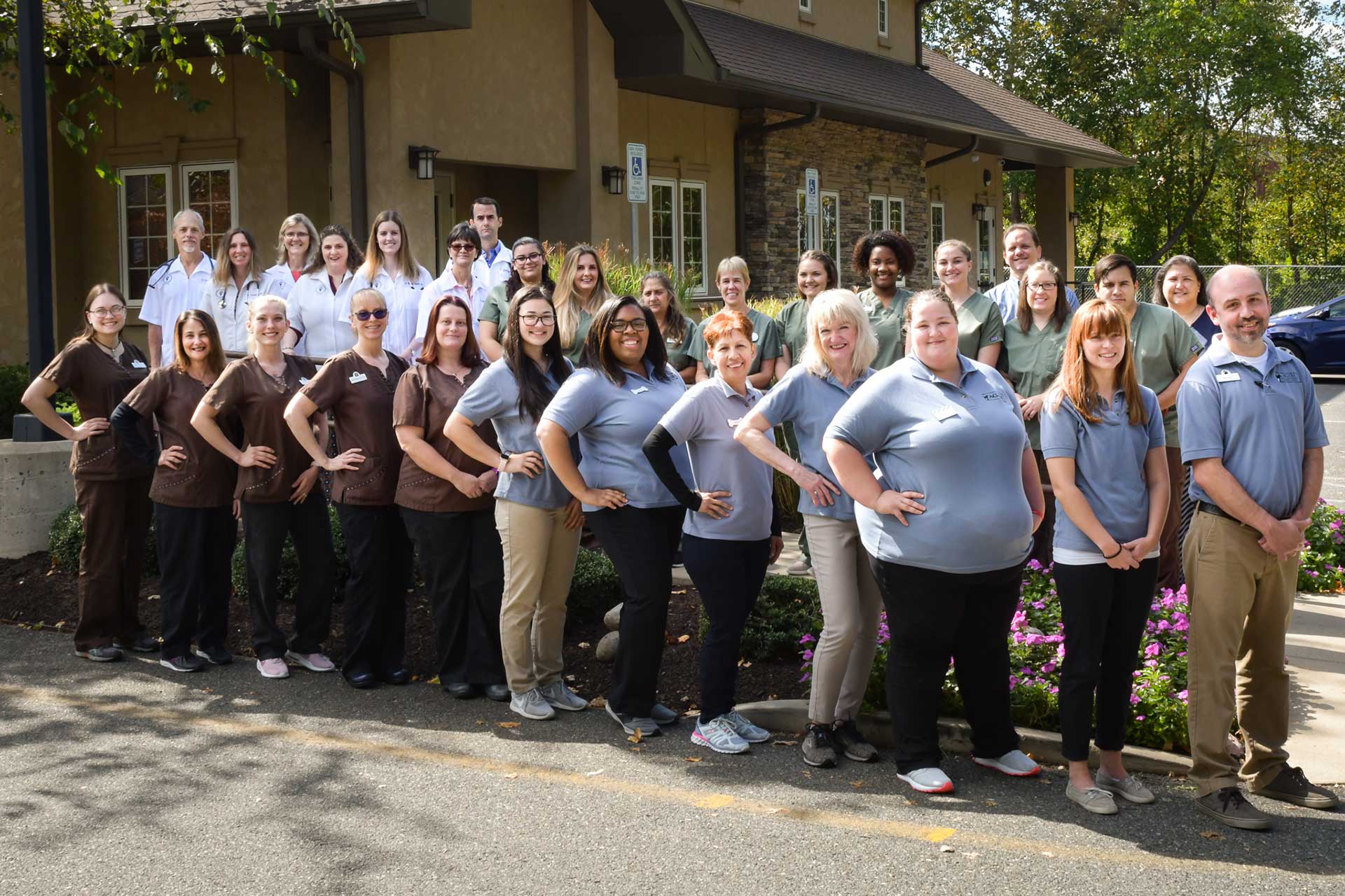A large group of people stands outside in front of a building, posing for a photo. They are dressed in various uniforms, some in brown and some in light blue. Most individuals, including several veterinarians, have their hands on their hips, and they are smiling at the camera.