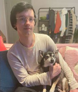 A person with glasses and dark hair, wearing a light-colored long-sleeve shirt, is sitting on a pink couch. They are holding a small black and gray dog like a caring vet might do. In the background, a clothing rack with various garments is visible.